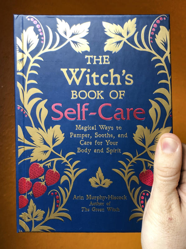 a photo of The Witch's Book of Self Care, with a dark blue cover featuring plant vines and fruit imagery